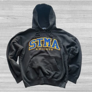 STMA Hoodie - Säljer en svart hoodie från Pennant med STMA Knights tryck i blått och gult på framsidan. Den har en klassisk känguruficka och justerbar huva med dragsko. Perfekt för en avslappnad stil.