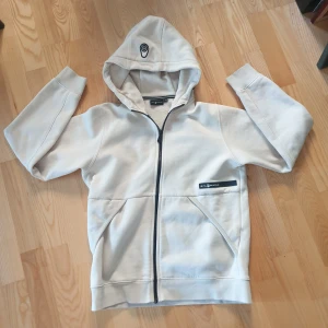 Vit hoodie från Sail Racing - Säljer en stilren vit hoodie från Sail Racing med dragkedja och huva. Tröjan har en logotyp på bröstet och en på huvan. Perfekt för en avslappnad stil.
