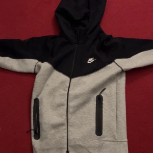 Grå och svart huvtröja från Nike tech - Säljer en stilren grå och svart huvtröja från Nike. Tröjan har en dragkedja framtill och två praktiska fickor med dragkedjor. Perfekt för en avslappnad stil. Den är helt ny och har bara använts en gång jag säljer på grund av att jag köpte en annan färg.