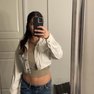 Vit croppad jeansjacka - Säljer en snygg vit croppad jeansjacka med knappar framtill. Jackan har en klassisk krage och långa ärmar. Perfekt för en trendig look!