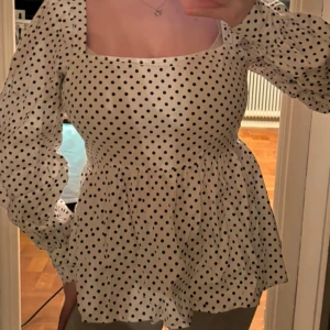 Vit prickig blus med puffärmar - Snygg vit blus med svarta prickar och puffiga långa ärmar. Den har en fyrkantig halsringning och en lätt utsvängd nederdel som ger en feminin touch. Perfekt för en stilren look!