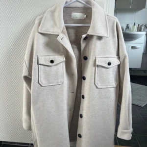 Beige jacka från Nelly - Säljer en stilren beige jacka från Nelly. Jackan har långa ärmar, knappar framtill och två stora fickor. Den är tillverkad i ett mjukt material och har ett bälte i midjan för en snygg passform. Är strl 34, men är oversize så passar S/M. Har någon mindre fläck men inte som syns så🥰