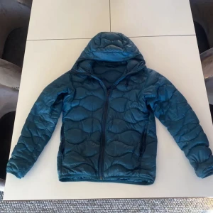 Blå dunjacka från Peak Performance - Säljer en snygg blå dunjacka från Peak Performance. Jackan har en quiltad design med dragkedja framtill och långa ärmar. Perfekt för kyligare dagar.