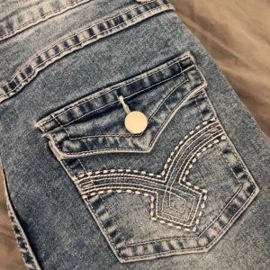 Blå jeans med broderad bakficka - Snygga lagom utsvängda blå jeans med en unik broderad bakficka och knappdetalj.  Storlek S (36)