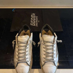 Vita sneakers från Golden Goose - Säljer ett par vita sneakers från Golden Goose med grå stjärndetalj på sidan och svarta hälpartier. Skorna har snörning och perforerade detaljer på tån. Perfekta för en stilren och avslappnad look.
