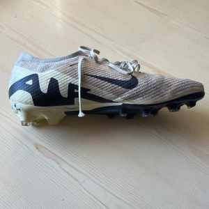 Nike fotbollsskor i beige och svart - Säljer bara vänstra fotens fotbollsskor i beige med svarta detaljer, eftersom den högra gick sönder. Skorna har snörning och en slitstark sula med dobbar för bra grepp på planen. Perfekta för fotbollsträning och matcher.