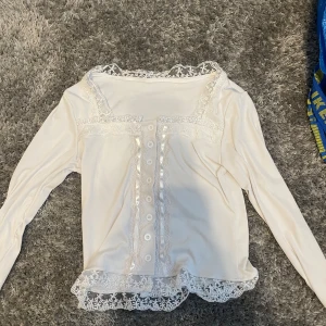 Vit långärmad topp med spetsdetaljer - Säljer en söt vit långärmad topp med knappar framtill och vackra spetsdetaljer vid halsringning och nederkant. Perfekt för en romantisk look. Passar bra till både jeans och kjol. Storlek: S