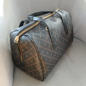 By Malene Birger  - Mycket fint skick, som ny.  Snygg väska med ett elegant mönster i brunt och svart. Väskan har en praktisk dragkedja i guld och bekväma handtag. Perfekt för att bära med sig det nödvändigaste med stil.