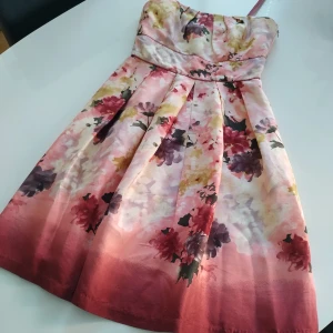 Blommig axelbandslös klänning från S. T. I. - Finskt mode. En vacker axelbandslös klänning från S. T. I i SIDEN med ett blommigt mönster i rosa, lila och röda toner. Klänningen har en elegant passform och är perfekt för sommarens festligheter. 🌸