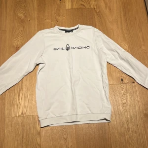 Vit tröja från Sail Racing - Säljer en stilren vit tröja från Sail Racing med svart text på bröstet. Tröjan har långa ärmar och en rund halsringning. Perfekt för en avslappnad stil.