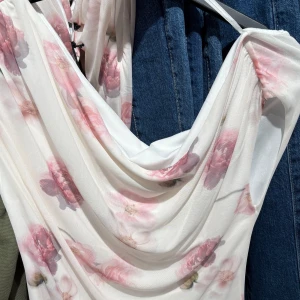 Vit blommig top  - Säljer en vacker vit klänning med rosa blommönster. Klänningen har en draperad design som ger en elegant look. Perfekt för somriga tillfällen.