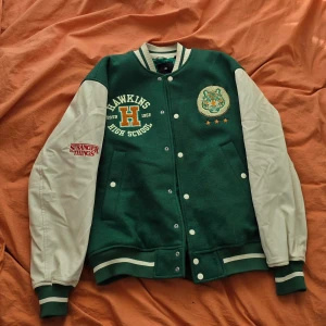 Stranger things collage jacka limited edition  - Säljer en grön och vit jacka med Hawkins High School tema från Stranger Things. Jackan har långa vita ärmar och grönt kroppsparti med tryck på både fram- och baksida. Perfekt för fans av serien!