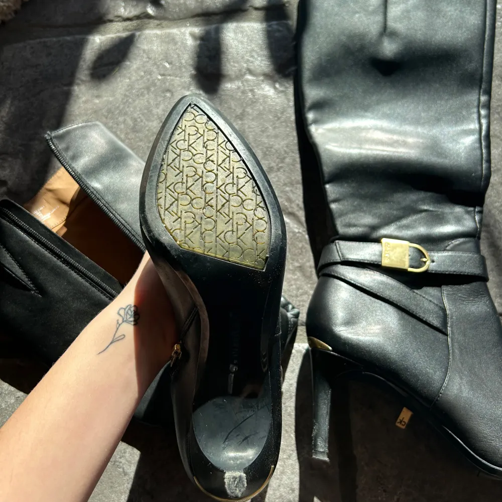 Snygga svarta stövlar i skinn med spetsig tå och klack. De har en dekorativ guldfärgad spänne och dragkedja på sidan för enkel på- och avtagning. Perfekta för en elegant look.. Kengät.
