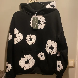 Svart hoodie med vita mönster från Denim Tears - Säljer en cool svart hoodie från Denim Tears med unika vita mönster över hela tröjan. Den har en klassisk känguruficka och en bekväm huva. Perfekt för att sticka ut i mängden!