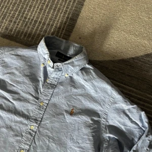 Blå skjorta från Ralph Lauren - Säljer en klassisk blå skjorta från Ralph Lauren med broderad logotyp på bröstet. Skjortan har en button-down krage och knappar framtill. Perfekt för en stilren look. Inga fel och 100% autentisk.