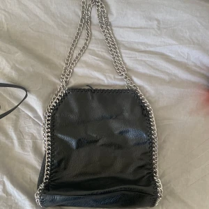 Svart axelväska från Tiamo - Snygg svart axelväska från Tiamo med kedjedetaljer längs kanterna och axelremmen. Väskan har en dragkedja för säker förvaring och är perfekt för att ge en stilren touch till din outfit.