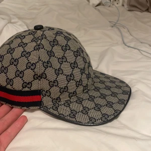 Gucci canvas grå navy blue  - Snygg keps med ett klassiskt mönster och logotyp. Den har en justerbar rem baktill och en randig detalj i rött och blått på sidan. Perfekt för att ge din outfit en stilfull touch.