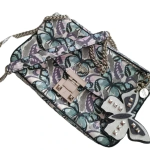 Färgglad axelväska från Guess - Snygg axelväska från Guess med fjärilsmönster i lila, blå och gröna nyanser. Väskan har en kedja som axelrem och ett dekorativt fjärilshänge med nitar. Perfekt för att ge din outfit en färgglad touch! (Som ny!, etiketten saknas)