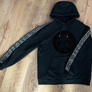 Svart Glorious Gangsta hoodie - Svart hoodie från Glorious Gangsta med stort svart broderat emblem på bröstet och feta mönstrade band längs ärmarna. Klassisk känguruficka framtill, snörning i huvan och riktigt skön passform. Perfekt för dig som gillar streetstyle. Priset kan diskuteras.