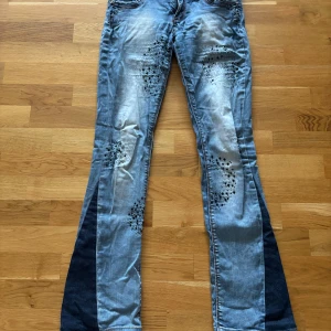 Blå bootcut jeans med coola detaljer - Unika blå jeans med bootcut och mörkare paneler längs benen. Snygga slitningar och svarta nitar på fram- och bakfickor ger en edgy vibe. Står ingen storlek men dom passar mig och jag har i vanliga fall 38 eller 29 i jeans.