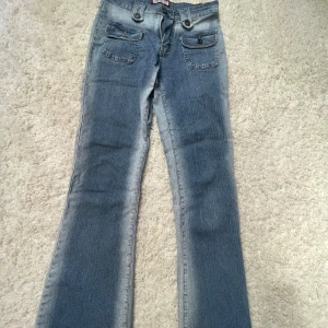 Blå bootcut jeans med fickdetaljer - Snygga blå bootcut jeans från Cross Jeans med två stora fickor framtill med lock och knappar. Jeansen har en klassisk tvättad look och är tillverkade i jeansmaterial. Perfekta för dig som gillar retrovibbar och coola detaljer.