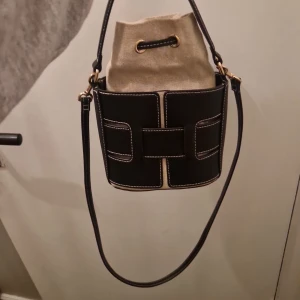 Svart och beige bucket bag med snörning - Cool bucket bag i svart och beige med kontrastsömmar och snygga detaljer i guld. Väskan har en unik design med svart ytterdel i skinn och beige innerpåse i tyg som stängs med snörning. Perfekt axelrem för att bära crossbody eller på axeln.