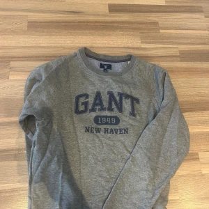 Grå sweatshirt från GANT XS - Snygg grå sweatshirt från GANT i storlek XS med blå text 'GANT 1949 NEW HAVEN' på bröstet. Klassisk rund halsringning och långa ärmar. Perfekt för en chill och stilren look.