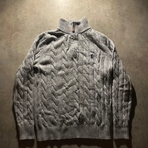 Grå kabelstickad tröja Polo Ralph Lauren - Kabelstickad grå tröja från Polo Ralph Lauren med half zip och hög krage. Klassisk design med ribbade muddar och broderad logga på bröstet. Perfekt för lager-på-lager och chill höst- eller vinterstil. Hör av vid minsta fundering 💫