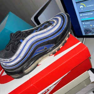 Nike Air Max 97 Black/Blue/Gold 42.5 - Nike Air Max 97 i svart med blå och silvriga vågformade detaljer samt guldigt swoosh. Ikonisk design med synlig Air-sula, snörning och mesh- och skinnpaneler. Perfekt för dig som vill sticka ut med en klassisk sneaker med modern vibe.