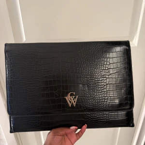 Svart datorfodral från CW - Stilren svart datorfodral från CW med croco-mönstrad skinnimitation och guldfärgad logga framtill. Insidan har ett snyggt mönster med CW-loggor och praktiska fack. Perfekt för att bära laptop eller viktiga papper med extra stil. 36x26 cm