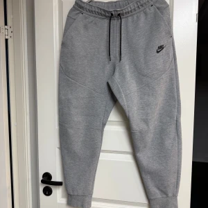 Grå Nike Tech Byxor - Snygga grå Nike Tech mjukisbyxor från Nike med svarta dragsnören i midjan och logga på vänster ben. Byxorna har ribbade muddar vid bensluten, praktiska fickor och en sportig look. Perfekta för chill eller träning.