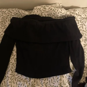 Svart offshoulder ribbad topp - Snygg svart offshoulder topp med ribbad struktur och långa ärmar. Perfekt för dig som vill ha en stilren och trendig look med en twist. Toppen har en bred kant upptill som ger en cool och modern känsla.