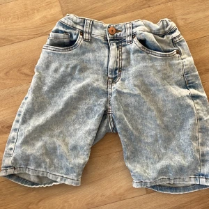 Ljusblå jeansshorts från Lindex - Snygga ljusblå jeansshorts från Lindex med klassisk femficksdesign och slitna detaljer. Shortsen har normal passform, bälteshällor och knappstängning. Perfekta för varma dagar och enkel att matcha med t-shirt eller hoodie.