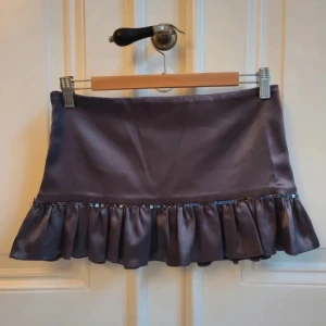 Grå satinminikjol med volang - Supersöt mörkgrå kort kjol i glansig satin med bred volangkant och små paljetter längs sömmen. Passar xs/s