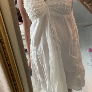 Vit somrig långklänning med smock - Luftig vit långklänning med smockad överdel och tunna axelband. Klänningen har en lätt volangkänsla upptill och knytband framtill. Perfekt för varma dagar och har en romantisk, bohemisk vibe. Vet ej märke men den är köpt utomlands och passar storlek XS-M