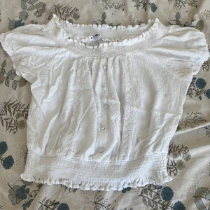 Vit blus med volang från FB Sister - Supersöt vit blus från FB Sister med korta ärmar, volangdetaljer vid hals och nederkant samt knappar framtill. Toppens tyg har diskret struktur och små prickar, och den har smockad midja för en snygg passform. Perfekt för dig som gillar romantisk stil.