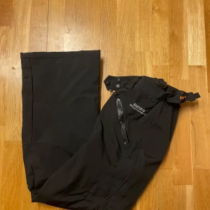 Svarta täckbyxor Ross Sportswear - Svarta täckbyxor från Ross Sportswear med justerbara hängslen, dragkedja och tryckknappar i midjan. Byxorna har vattentätt material och en dragkedjeförsedd ficka framtill. Perfekta för vinterns alla äventyr.