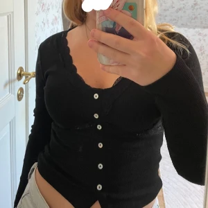 Svart kofta med spets från Gina Tricot - Supersnygg svart tröja från Gina Tricot med v-ringning och spetsdetaljer längs halsringningen och under bysten. Tröjan har ribbad struktur, knappar framtill och lång ärm. Perfekt för en snygg vardagsoutfit! Aldrig använd och har prislappen kvar, nypriset är 299kr. 