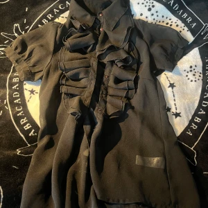 Svart volangskjorta från Sara Kelly - Svart skjorta från Sara Kelly x Ellos med korta ärmar och transparent tyg. Snygga volangdetaljer framtill och röda knappar som ger extra edge. Perfekt för dig som gillar statement-plagg och vill sticka ut lite extra.