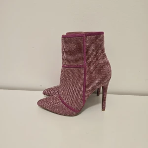 Glittriga rosa stövletter med klack - Snygga stövletter i glittrigt rosa material med spetsig tå och hög klack. Skorna har dragkedja bak och är täckta av små glittrande stenar som ger en riktigt festlig vibe. Perfekta för dig som vill sticka ut och addera extra bling till outfiten.