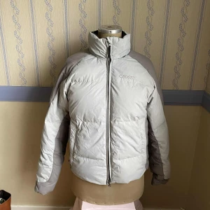 Ljusgrå pufferjacka - Ljusgrå pufferjacka från Cross med hög krage, svarta detaljer och dragkedja framtill. Dunjackan har luva som går att ta ut ur kragen. 🩷