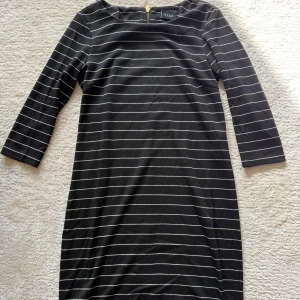Svart randig klänning från Vila XS - Svart kortklänning från Vila i storlek XS med tunna vita horisontella ränder. Klänningen har trekvartsärm och en snygg guldig dragkedja i ryggen. Den är figurnära och passar perfekt för en stilren look.