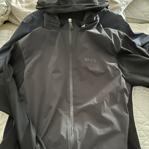 Svart vindjacka från BOSS med huva - Snygg svart vindjacka från BOSS med huva och hel dragkedja framtill. Jackan har stilren design med diskret BOSS-logga på bröstet och på dragkedjan. Perfekt för dig som gillar minimalistisk och sportig stil.