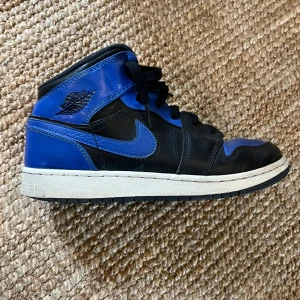Nike Air Jordan 1 Mid Svart/Blå - Säljer ett par Nike Air Jordan 1 Mid i svart och blått skinn med klassisk vit sula och svarta skosnören. Ikonisk Jordan-logga på sidan och perforerad tåbox för extra stil. Perfekta för dig som vill sticka ut med en tidlös sneaker.