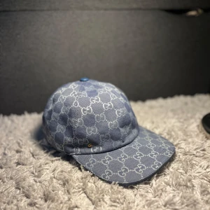 Blå Gucci keps - Snygg blå keps från Gucci med klassiskt GG-monogram över hela kepsen. Justerbar rem baktill och diskret guld-detalj på sidan. Tillverkad i bomullsmaterial med insida i beige tyg. Perfekt för dig som vill sticka ut med en lyxig accessoar.