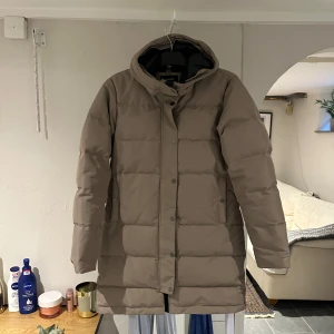 Beige pufferjacka från Everest - Säljer en beige pufferjacka från Everest med huva och dragkedja framtill. Jackan är lång, har knappar längs öppningen och två fickor med lock. Perfekt för kalla dagar och riktigt skön att bära. Ny