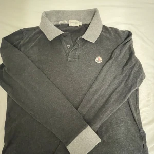 Grå långärmad pikétröja Moncler - Snygg långärmad pikétröja från Moncler i mörkgrått med ljusgrå krage och muddar. Tröjan har klassisk pikéknäppning med två knappar och Moncler-logga broderad på bröstet. Slim fit-modell i mjuk bomull, perfekt för en clean och stilren look.