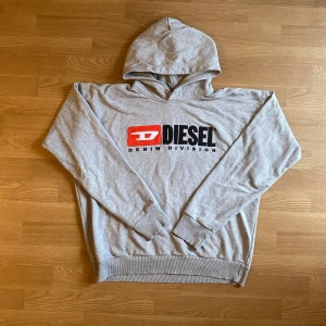 Grå Diesel hoodie - Säljer en grå hoodie från Diesel med stor broderad logga i svart och rött framtill. Tröjan har huva med metallöljetter, långa ärmar och ribbade muddar. Tillverkad i 100% bomull för en skön och avslappnad känsla. Nypris 1100kr