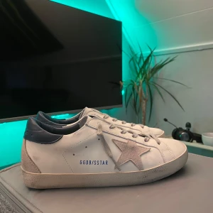 Golden Goose Superstar - Golden Goose Superstar sneakers i vitt skinn med grå mockadetaljer och blå hälkappa. Klassisk stjärna på sidan, grå snörning och platt sula. Snyggt slitna detaljer för en cool streetstyle-look. Kommer med originalkartong och dustbag.