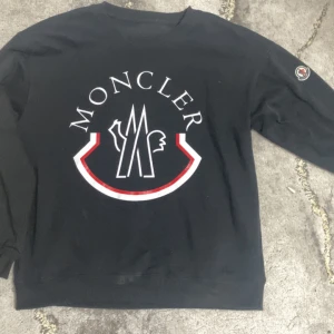 Moncler sweatshirt - Svart sweatshirt från Moncler med stort vitt och rött logotryck på bröstet och klassisk Moncler-patch på ärmen. Tröjan har rund hals och långa ärmar, perfekt för en clean och stilren look. Materialet känns mjukt och bekvämt, passar till många outfits.